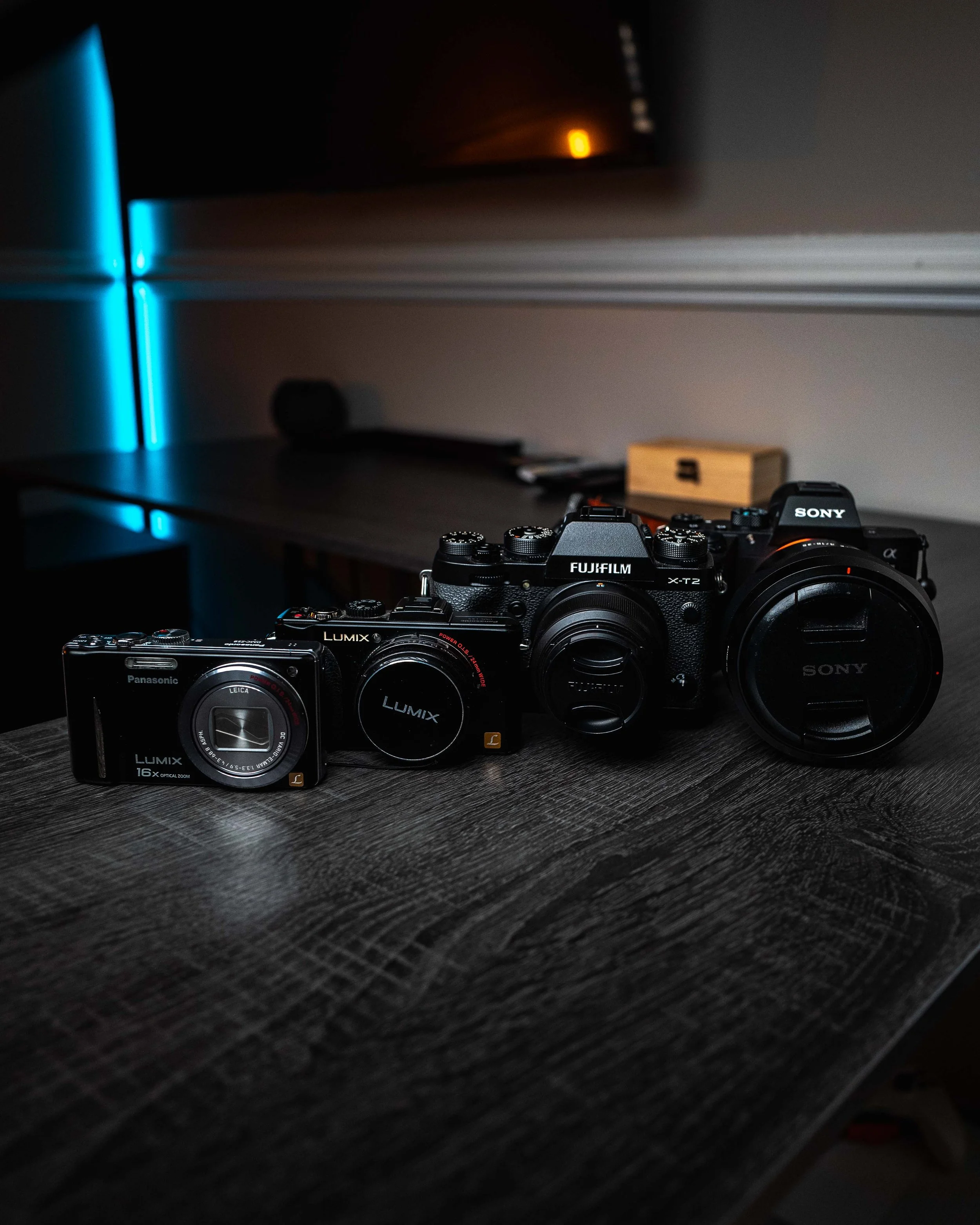 Three cameras placed on a dark wooden surface, with a dimly lit room and blue and orange lights in the background.