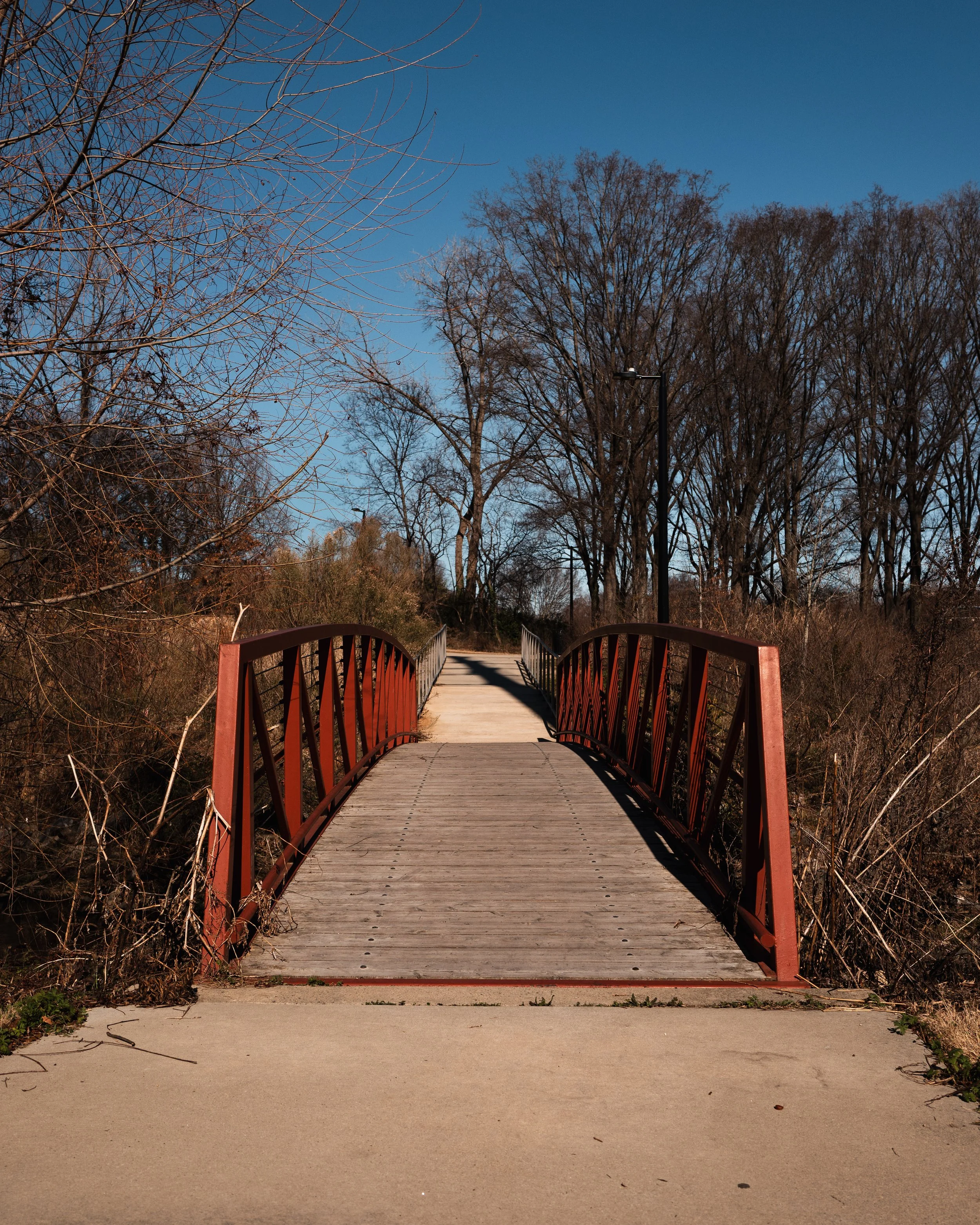 A small wooden pedestrian bridge with red metal railings over a stream, surrounded by leafless trees under a clear blue sky.