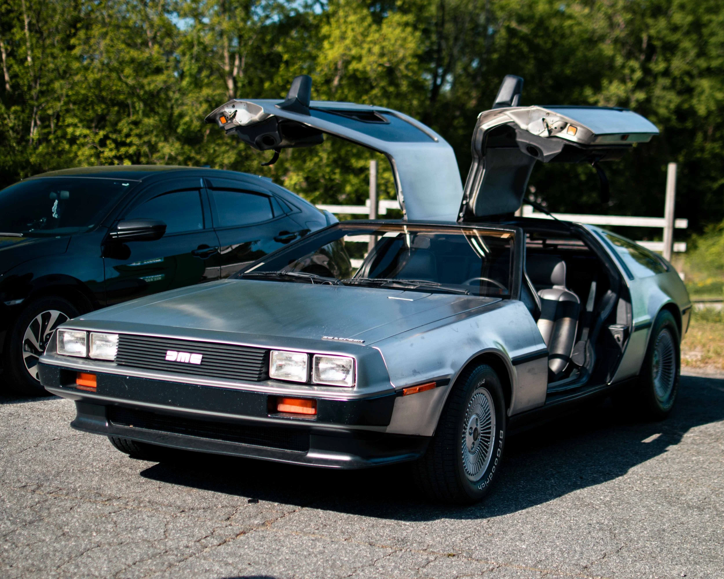 A DeLorean DMC-12 car with gull-wing doors open, parked on a road with trees in the background.