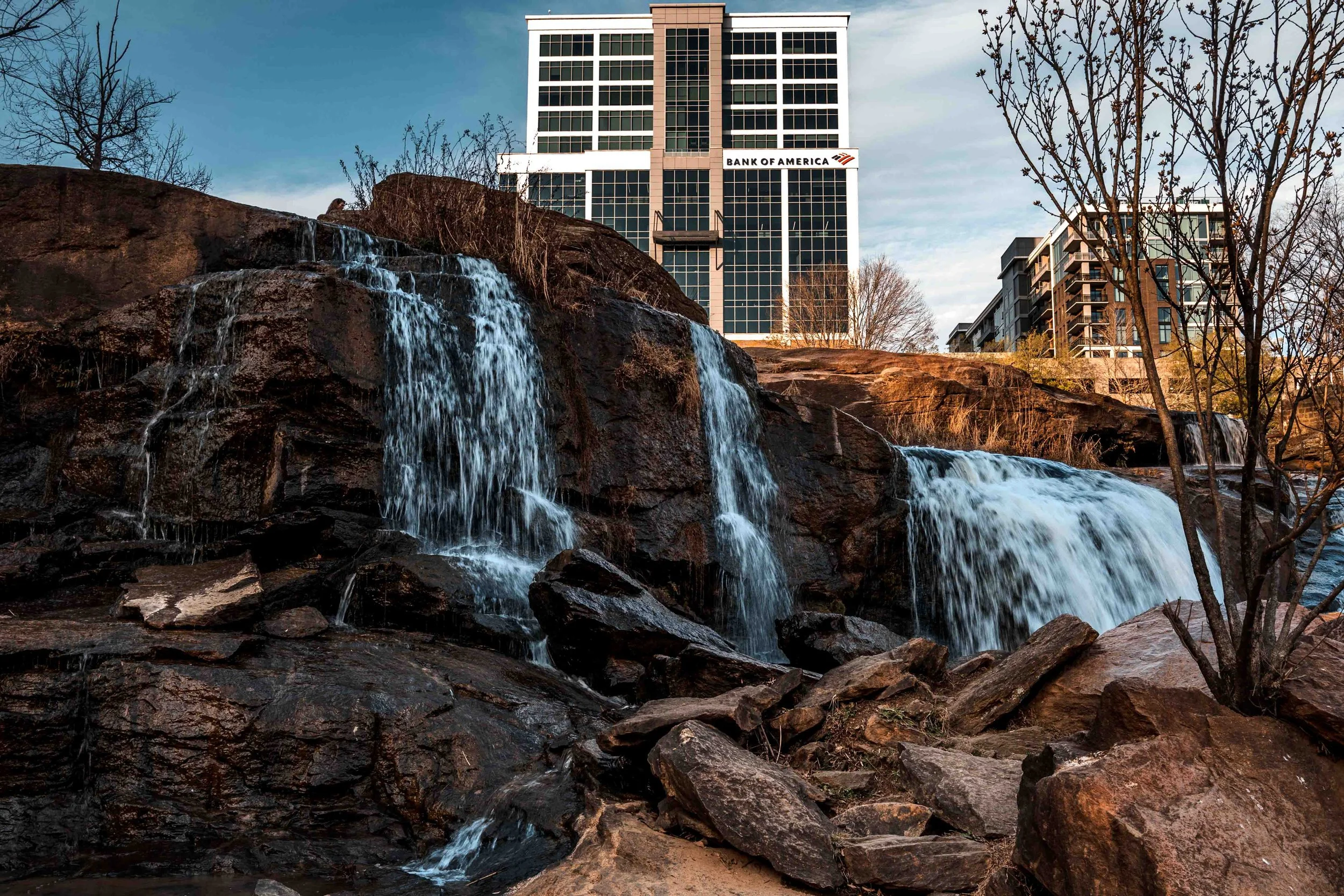 Waterfall cascading over rocks with a cityscape, including a tall building with the sign 'Bank of America,' in the background