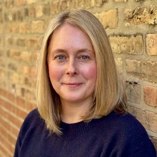 Woman with blonde hair wearing a dark sweater standing in front of a brick wall.