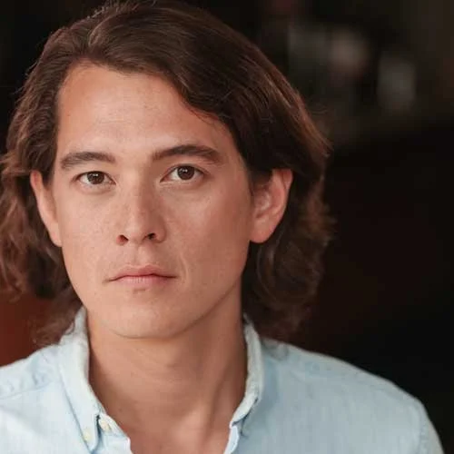 A young man with shoulder-length brown hair, wearing a light blue collared shirt, looking directly at the camera with a neutral expression.