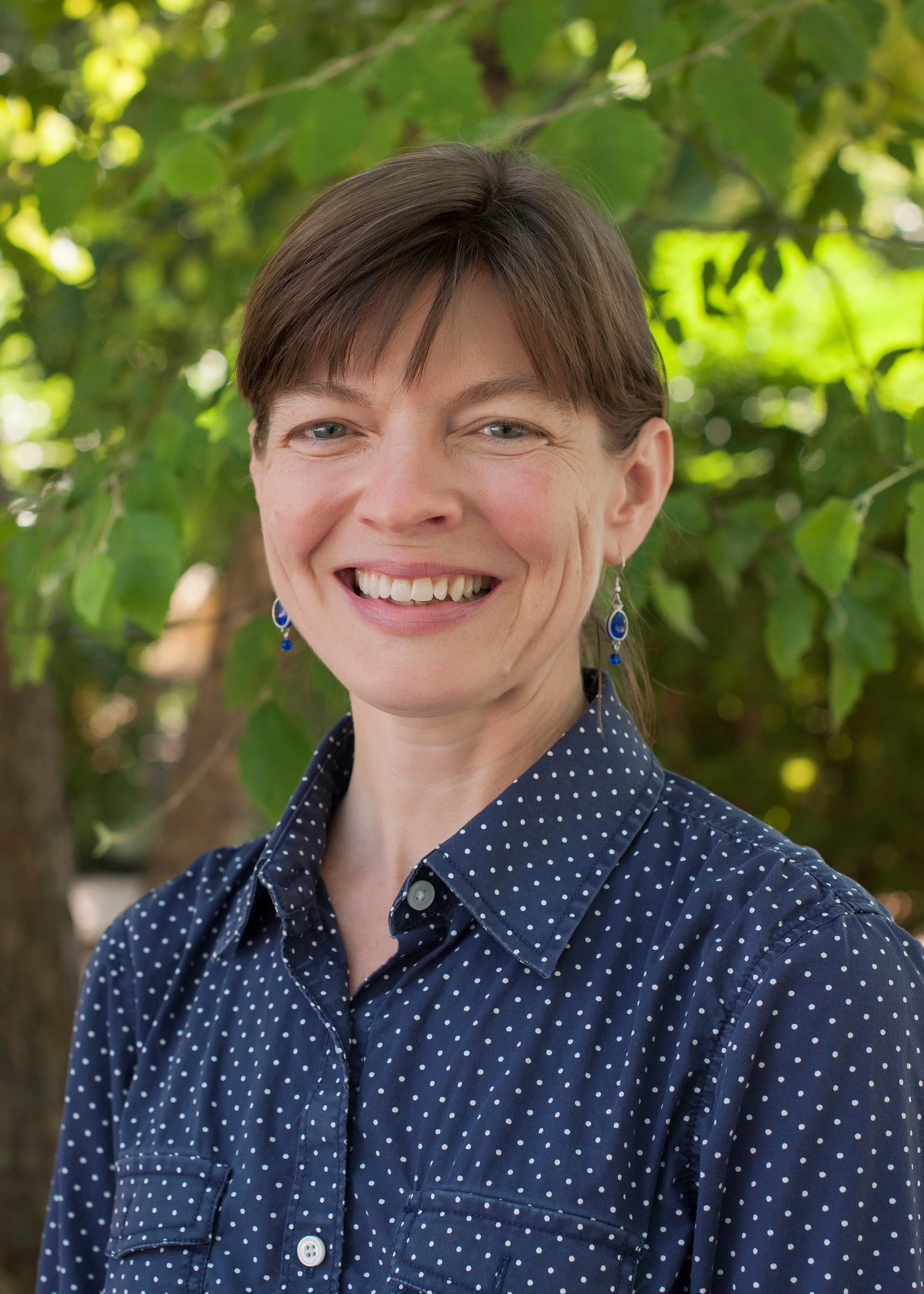 A woman with short brown hair and blue eyes smiling outdoors, wearing a navy blue polka dot shirt and blue earrings.