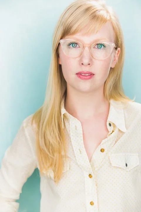 Woman with long blonde hair wearing glasses and a light-colored button-up shirt standing against a light blue background.