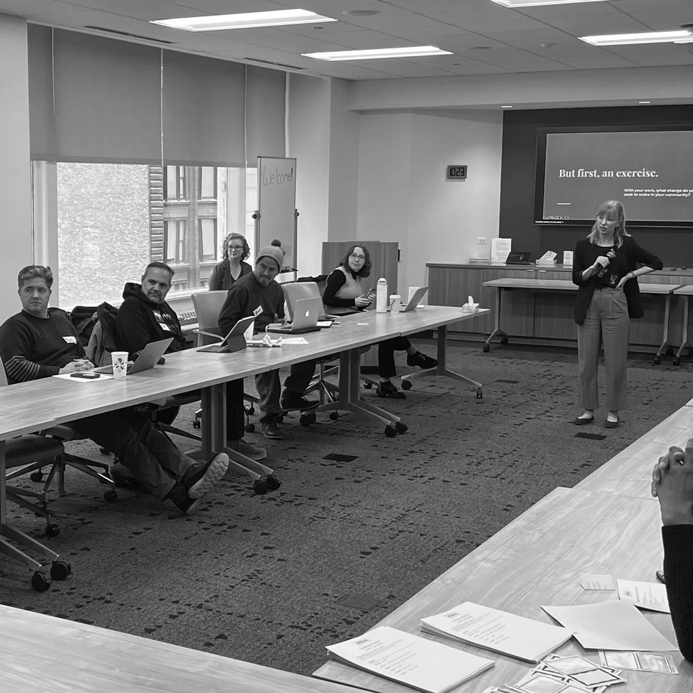 A woman is giving a presentation to a group of five seated individuals in a conference room, with a large screen and a whiteboard behind her.