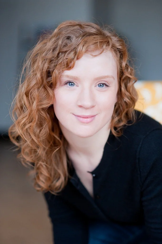 Close-up of a young woman with curly red hair and blue eyes, smiling softly, wearing a black top indoors.