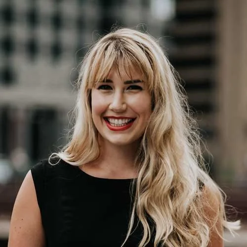 A smiling woman with long blonde hair, wearing a black top, standing outdoors with an urban background.