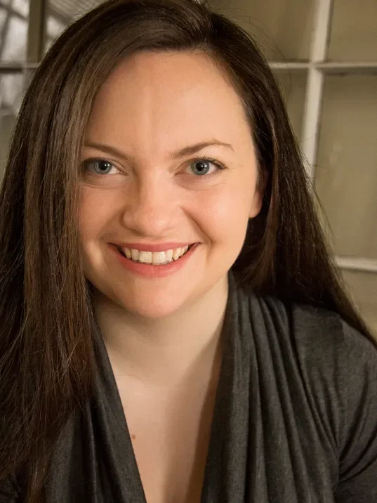 A woman with long brown hair and blue eyes smiling at the camera, wearing a gray top, with a blurred background behind her.
