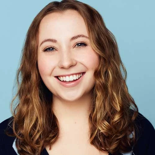 A young woman with wavy light brown hair smiling against a light blue background.