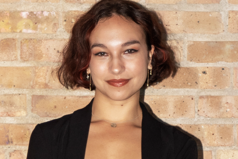 Portrait of a young woman with short wavy brown hair, wearing a black top, gold earrings, and a delicate necklace, standing against a brick wall.