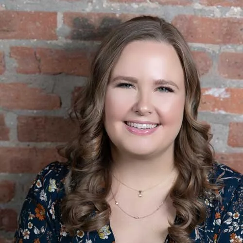 A young woman smiling in front of a brick wall, with brown curly hair and wearing a floral top and layered necklaces.