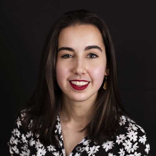 A young woman with long dark hair, wearing a black and white floral top, smiling at the camera against a dark background.