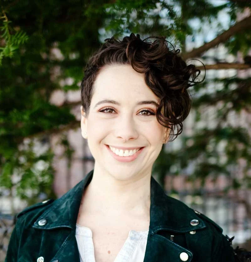 A smiling woman with short, curly dark hair wearing a leather jacket standing outdoors with green trees in the background.