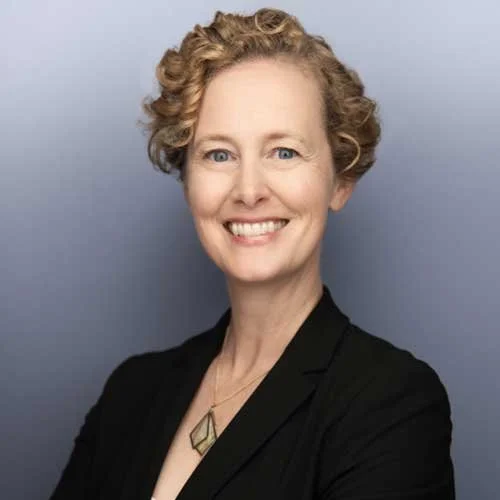 A woman with short curly blonde hair, blue eyes, and fair skin, smiling and wearing a black blazer and a geometric necklace, against a light grey background.