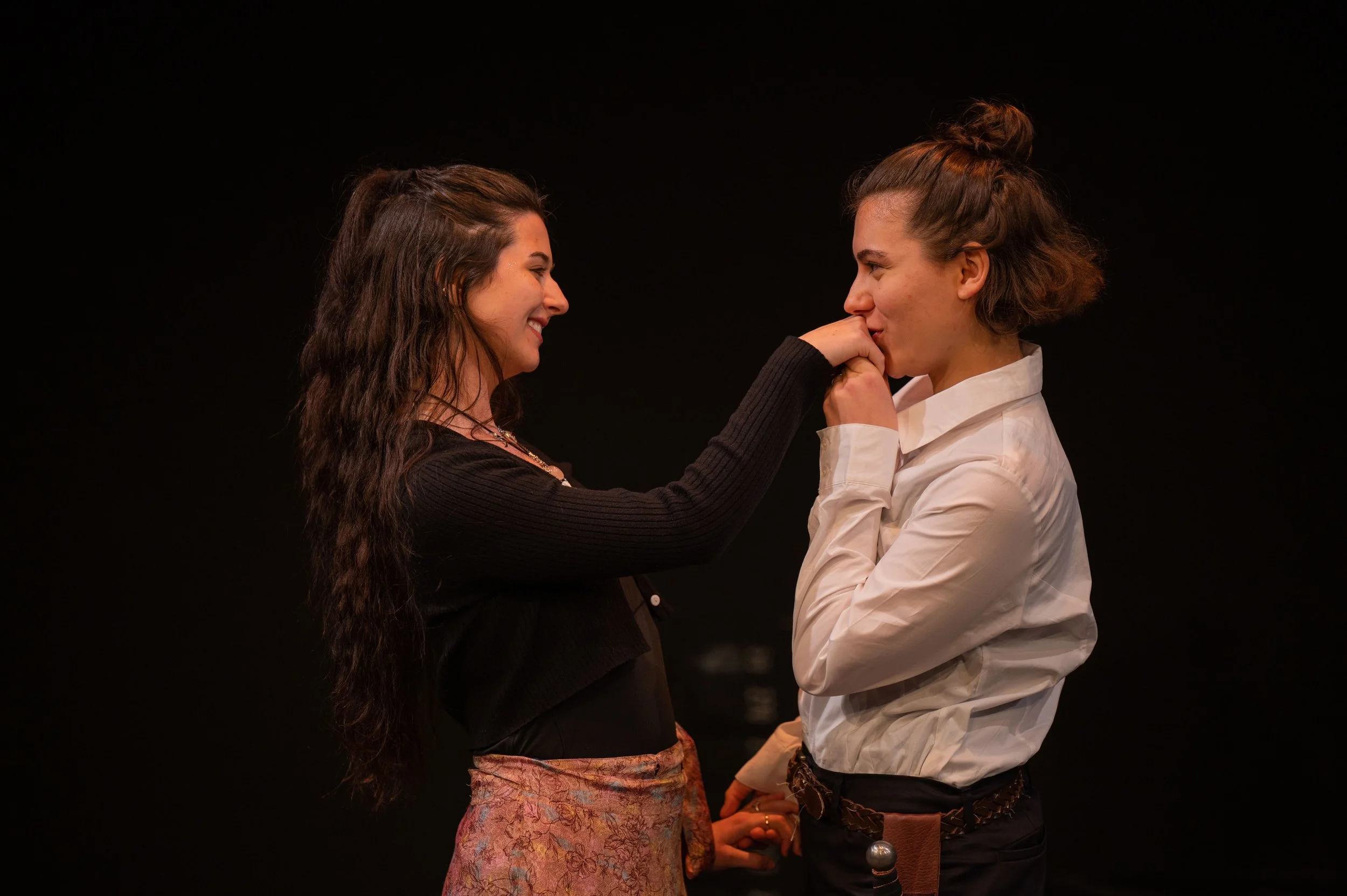 Two women holding hands and smiling at each other against a black background.