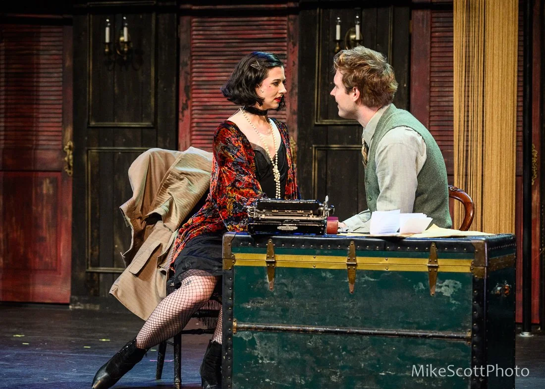 A woman with black hair and vintage-style clothing sitting at a desk facing a man with blond hair in a green vest and white shirt. They are in a room with dark wooden walls and a closed door in the background.