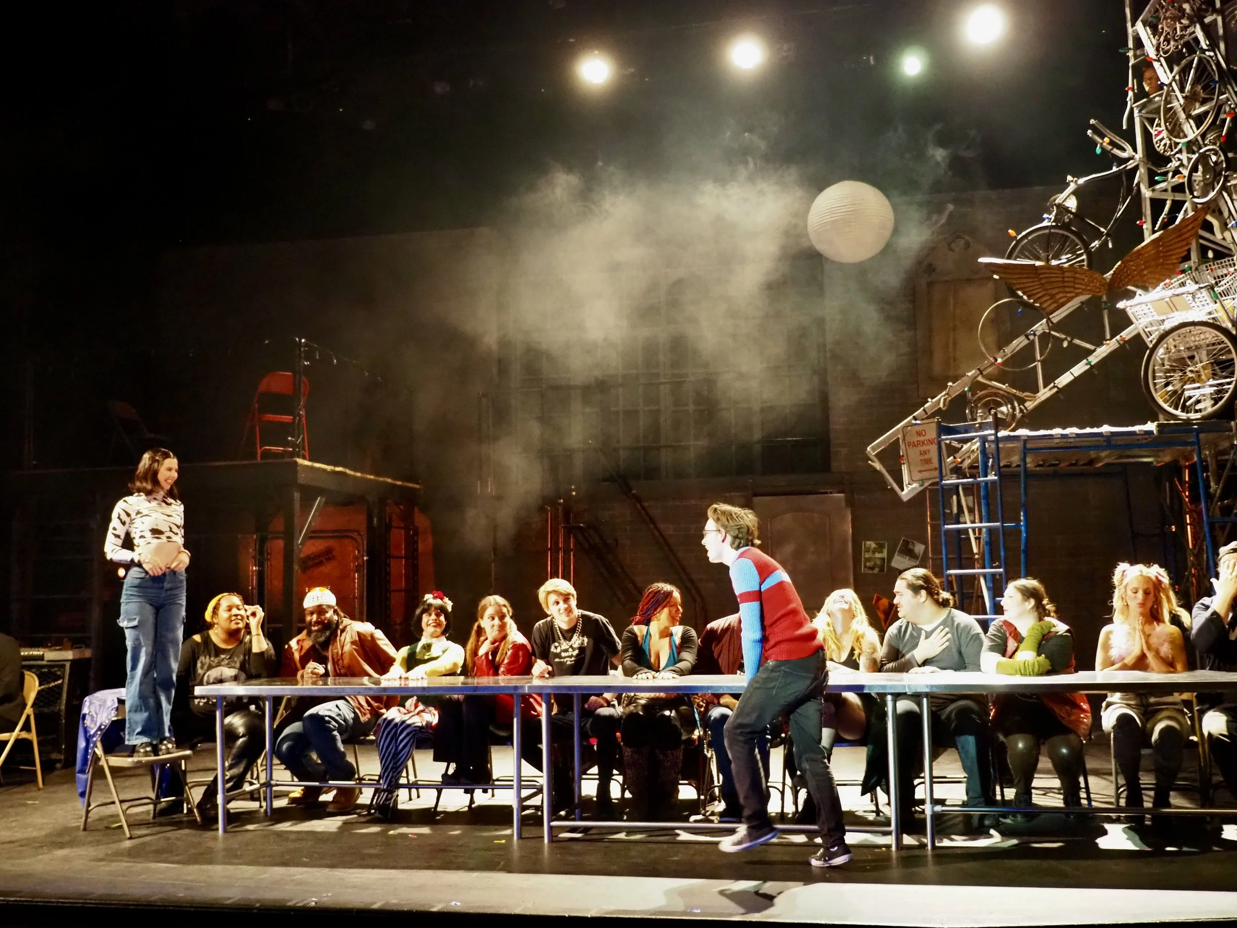 People in a theater rehearsal or performance scene, with a woman standing on a chair and a man dancing in front of a long table, surrounded by other seated individuals, stage lighting, and stage props including bicycles and a large winged wheel.
