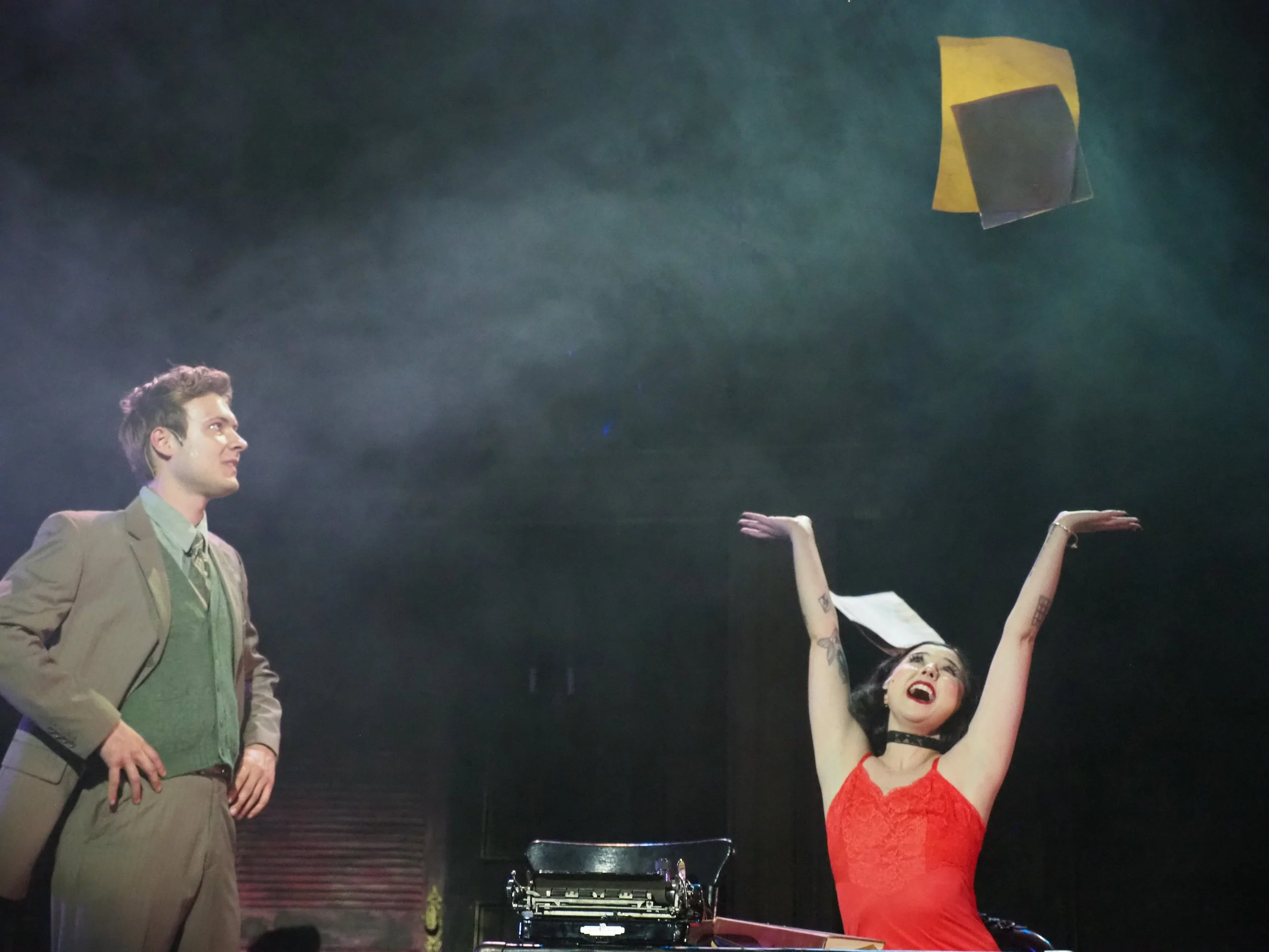 A man in a suit looks at a woman in a red dress who is throwing papers into the air and cheering on a stage with a typewriter and some objects.