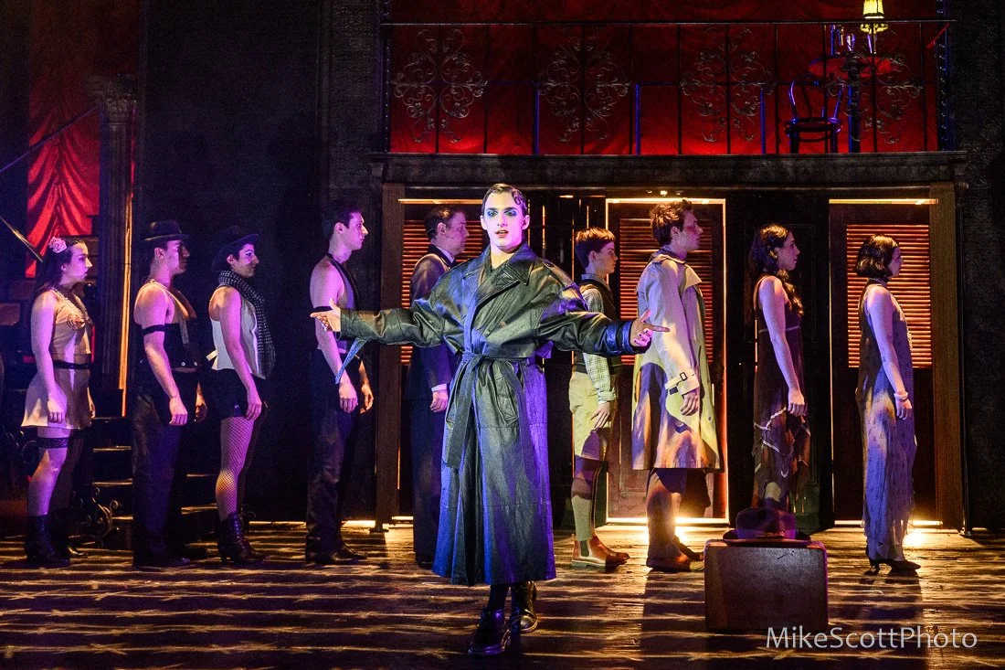 Stage scene with performers, featuring a woman in a black trench coat at the center, two levels of performers in vintage-style costumes, illuminated with dramatic lighting.