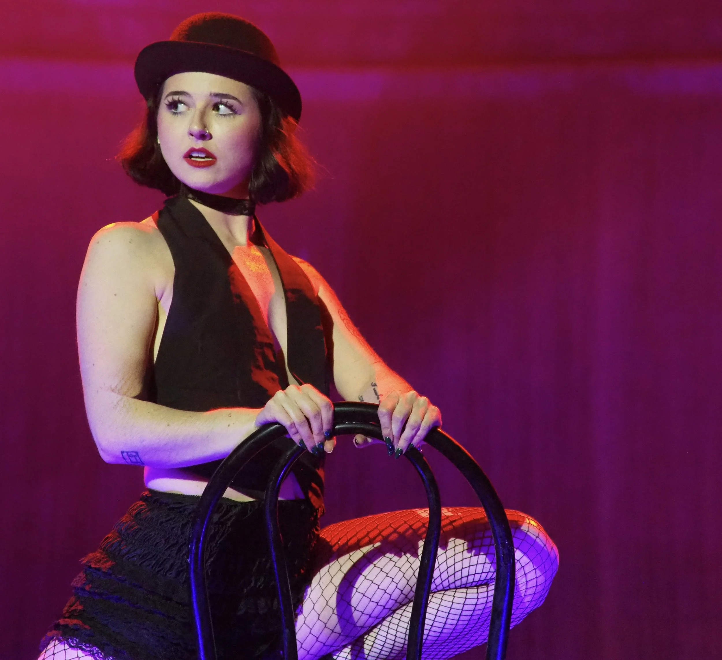 A young woman with short dark hair, wearing a black hat, sleeveless black top, fishnet tights, and black shorts, is holding onto a black chair while kneeling on one knee against a purple and pink gradient background.