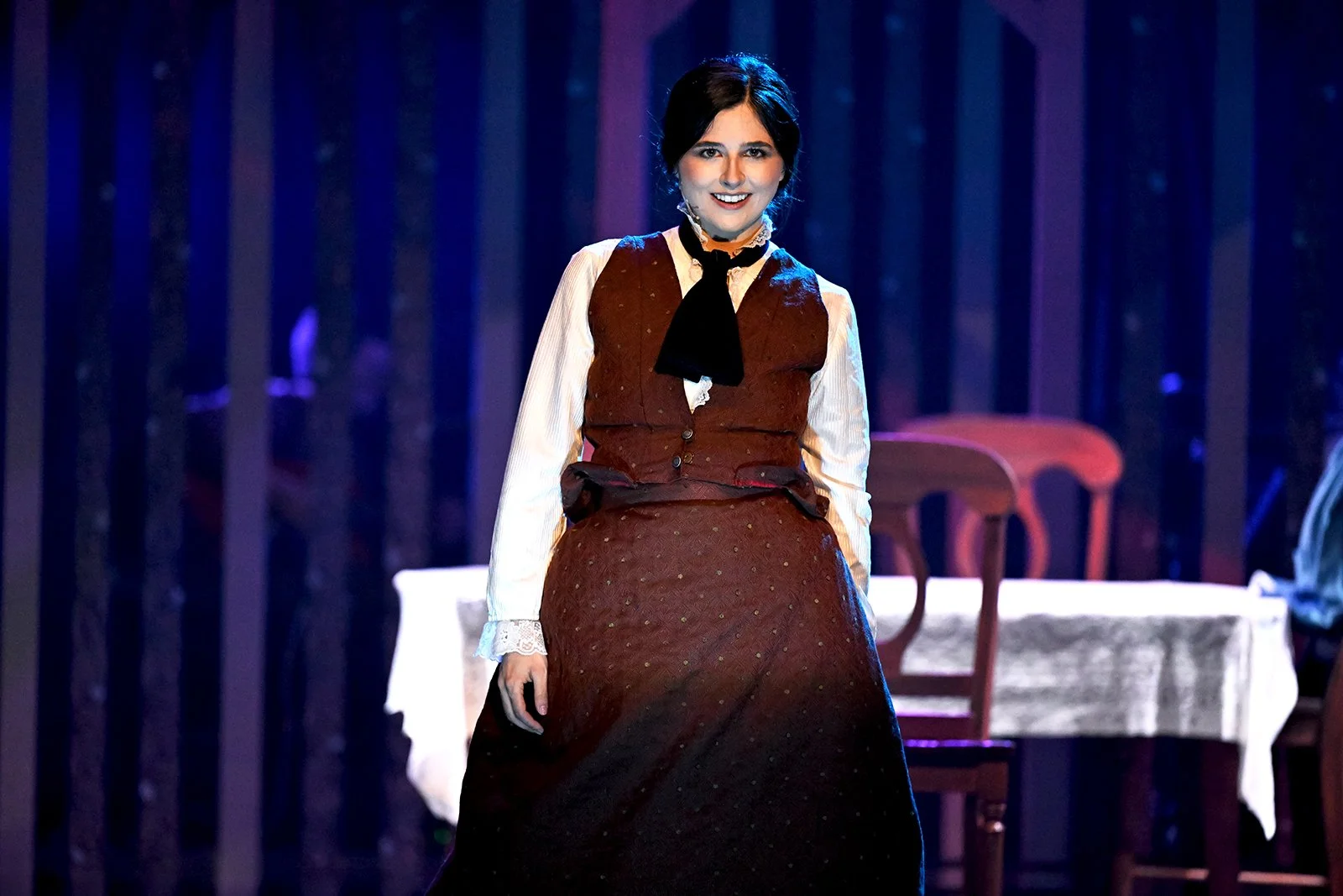 A woman dressed in period costume standing on stage with a table and chairs behind her, in a theater setting.