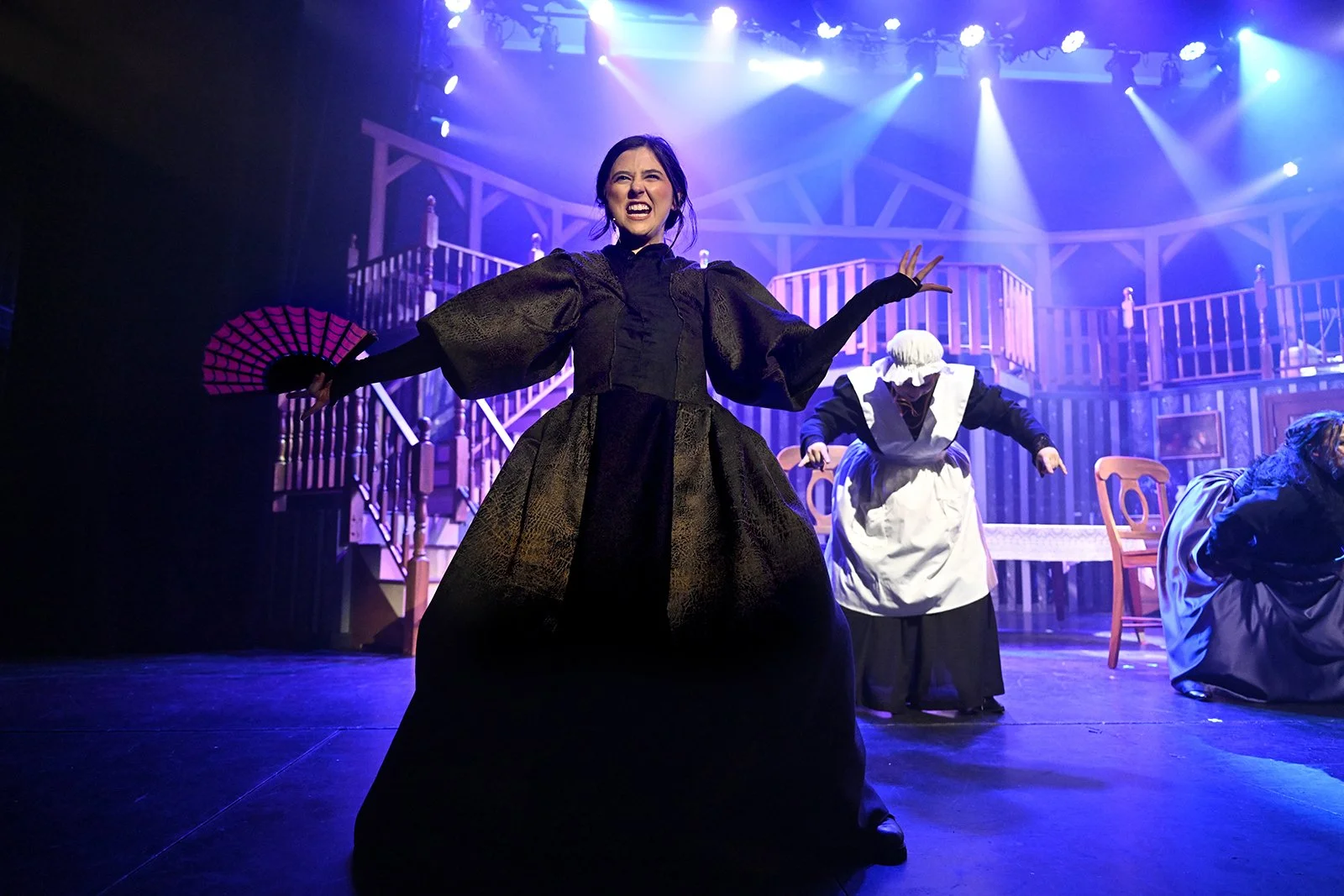 Performance on stage with women in historical costumes, woman in foreground holding a fan, stage with wooden furniture, set design resembling a house interior, dramatic lighting.