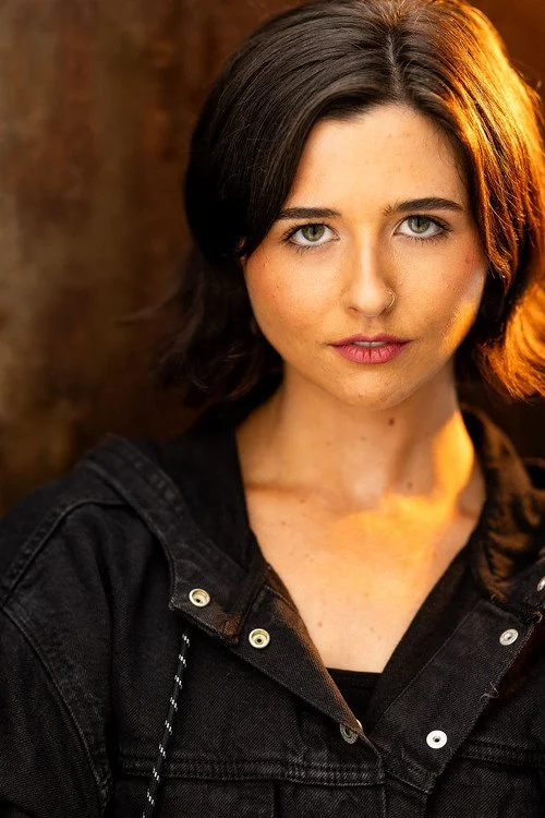 A young woman with short dark hair and a nose piercing, wearing a black jacket, posing outdoors in warm lighting.
