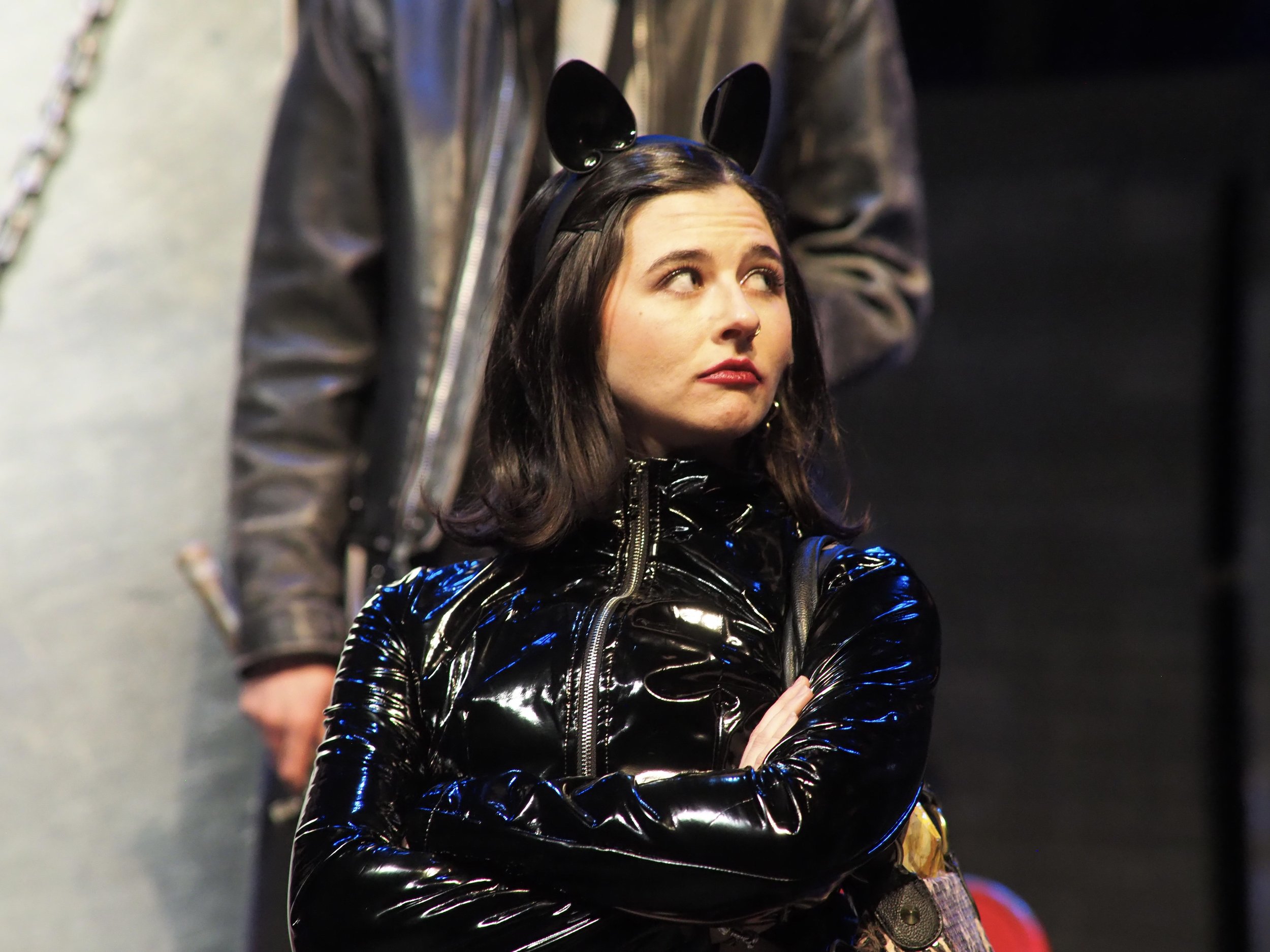 A woman with black hair in a black latex jacket and bunny ears headband looks to the side with crossed arms, standing against a dark background.