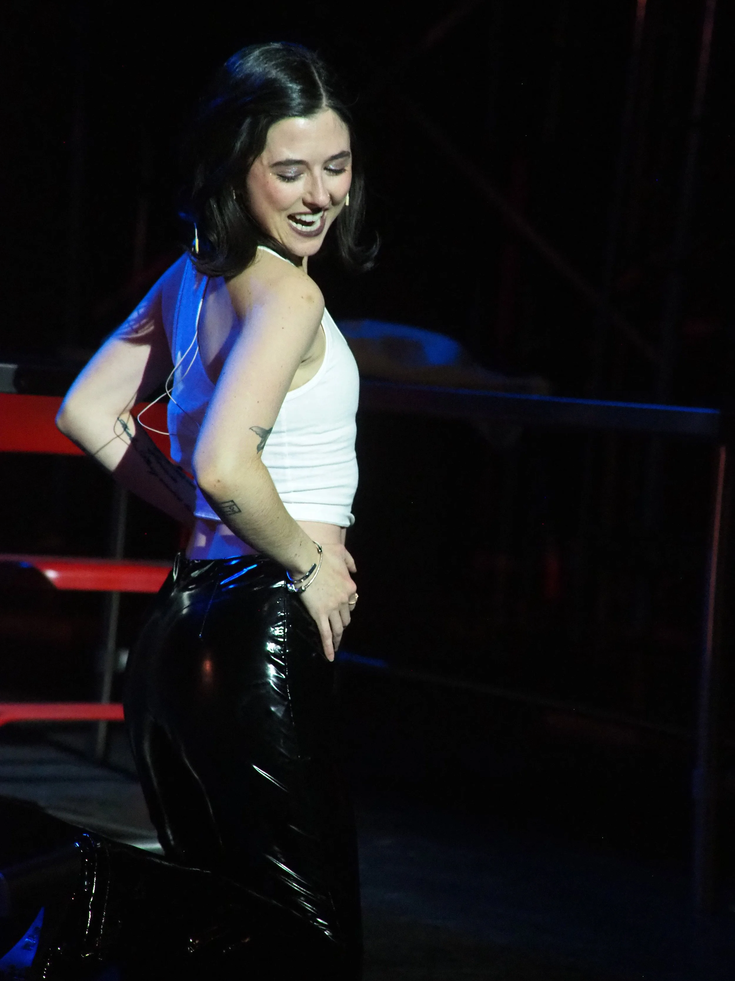 A woman with black hair, wearing a white tank top and shiny black pants, is smiling on a dark stage with red chairs in the background.