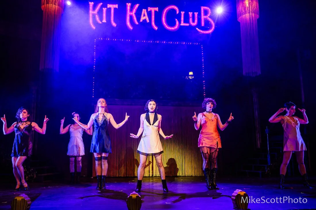 A stage performance featuring six women in 1920s-inspired costumes, with the sign 'KIT KAT CLUB' illuminated above them in pink and purple lights
