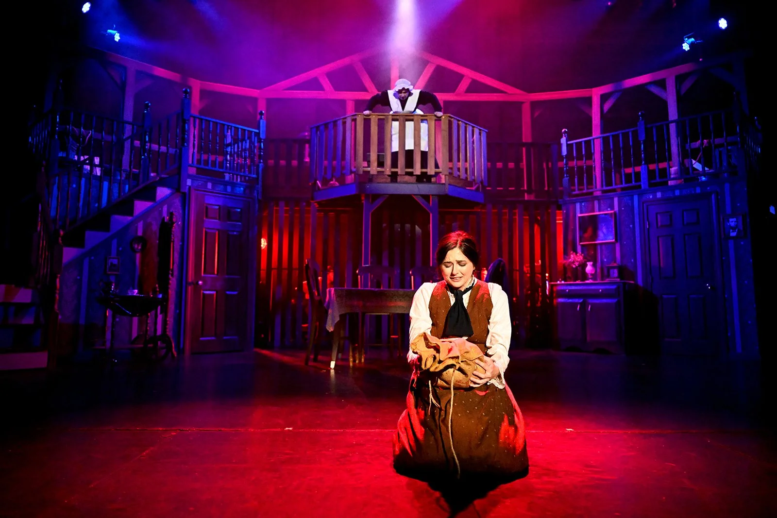 Woman dressed in historical costume kneeling on stage with a distressed expression, holding a bundle, in front of a two-story set resembling a house with purple and red lighting.
