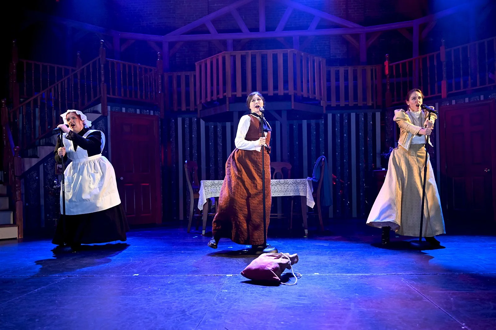 Three women on stage performing a play, dressed in period costumes, with a table and chairs in the background, and a bag on the floor in front.