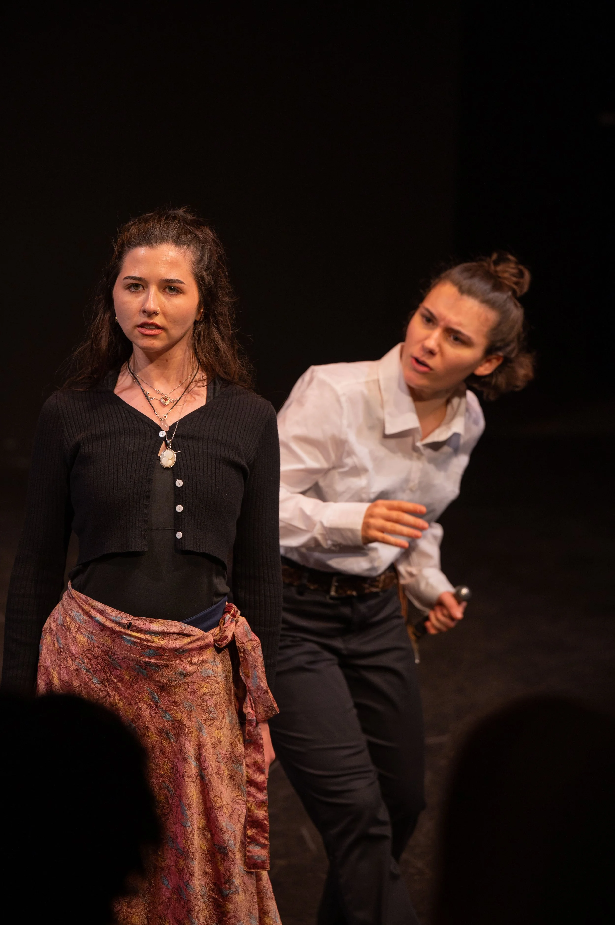 Two women performing on stage. One woman in front with dark hair, black top, and patterned skirt. The other woman behind, with brown hair tied up, wearing a white shirt, and holding a microphone.