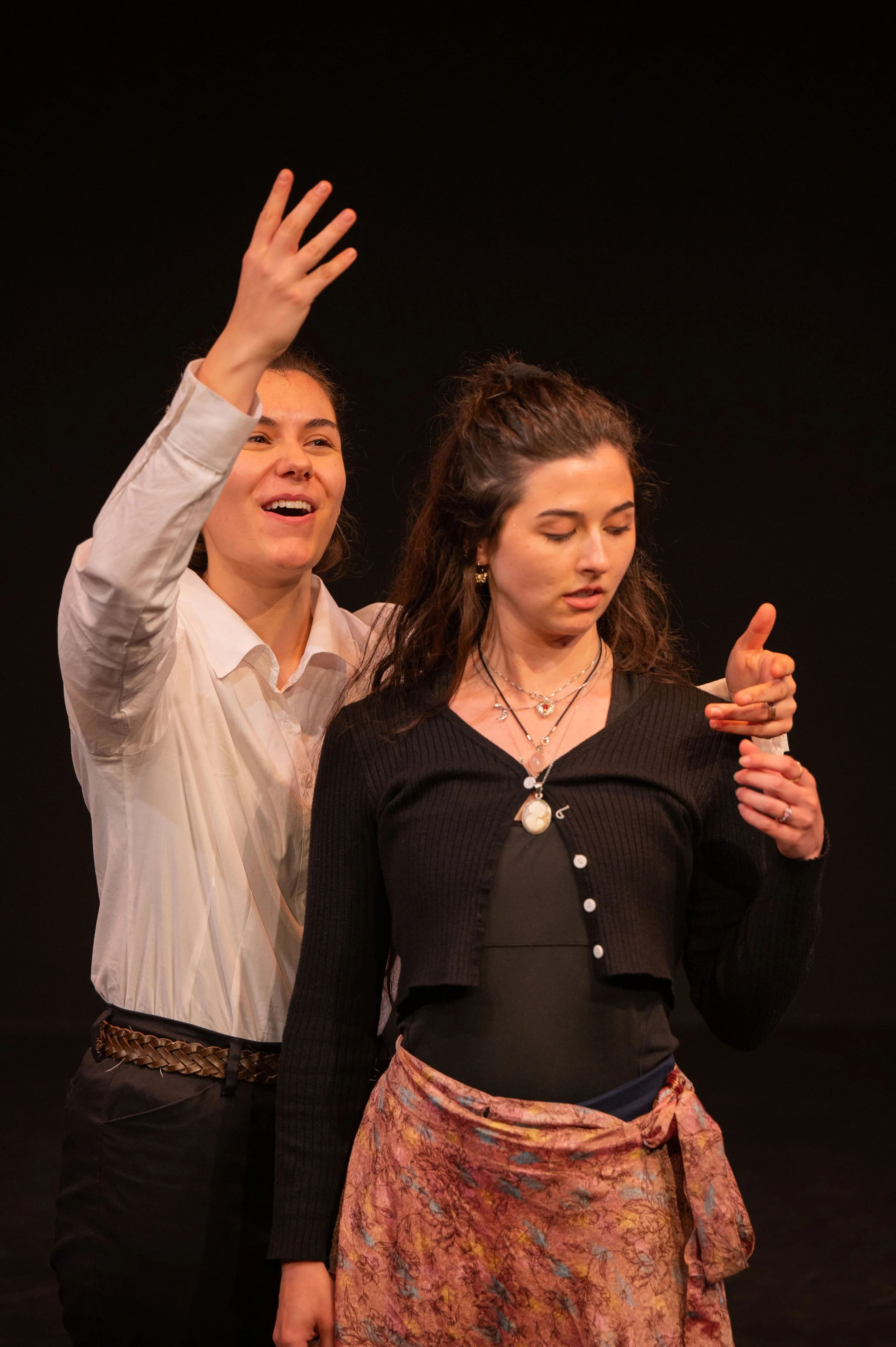 Two women on stage, one smiling and waving, the other looking down, both dressed casually, against a black background.