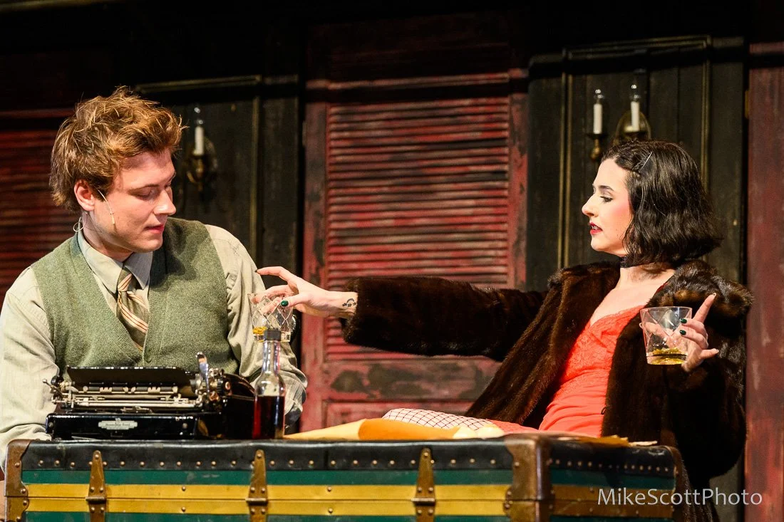 A man and woman sitting at a desk in a dramatic scene, with the woman extending her arm towards the man, who is looking down. The woman has dark, wavy hair, is wearing a fur coat and red dress, holding a glass of whiskey. The man has light brown hair, a vintage style, and is dressed in a vest and tie. The desk has a typewriter and bottles of alcohol, with a rustic background.