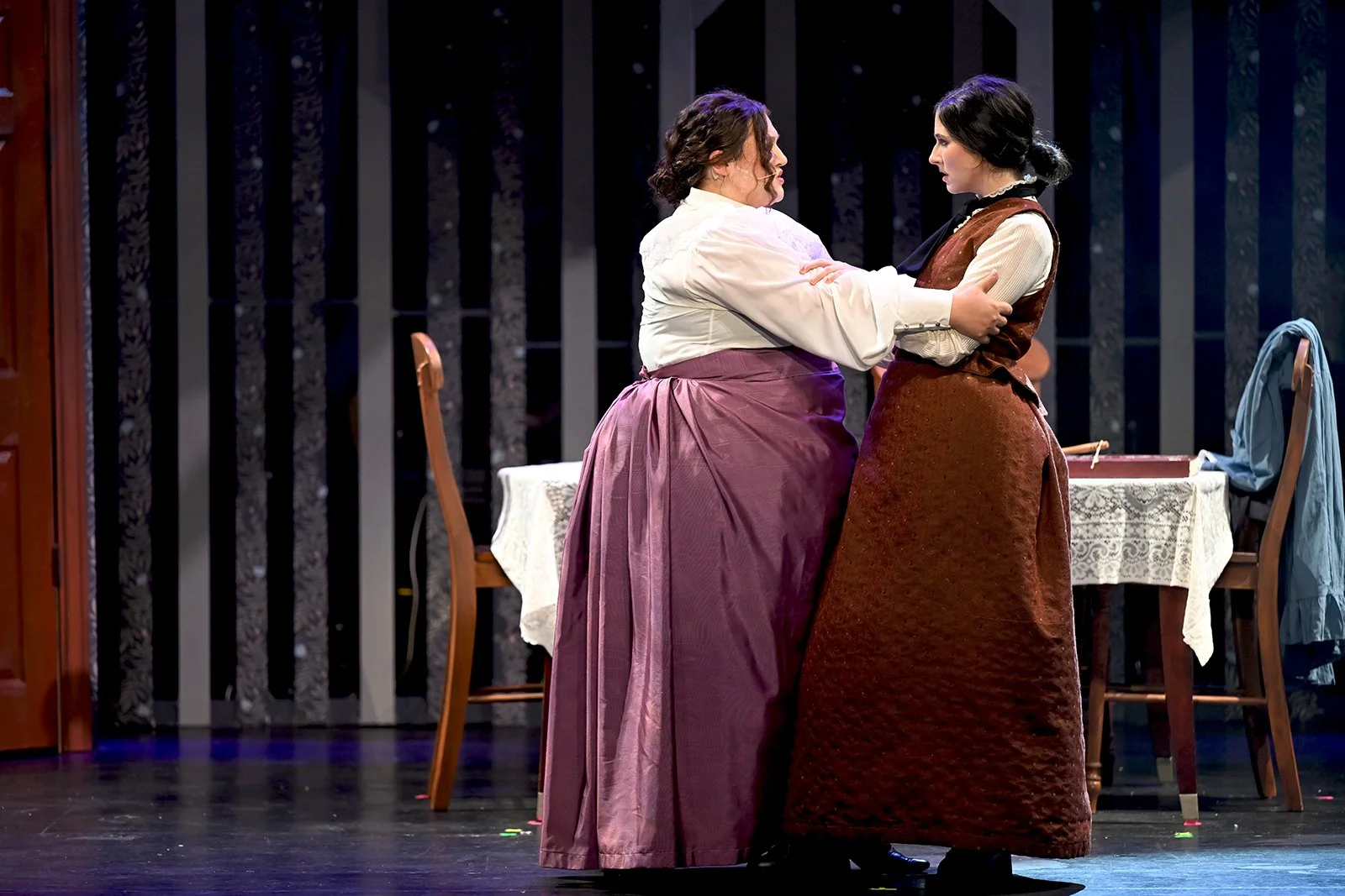 Two women dressed in historical clothing stand close together on a stage, engaging in a dramatic confrontation. A table with a lace tablecloth and a chair are visible behind them, amidst a dark backdrop with vertical wooden beams or panels.