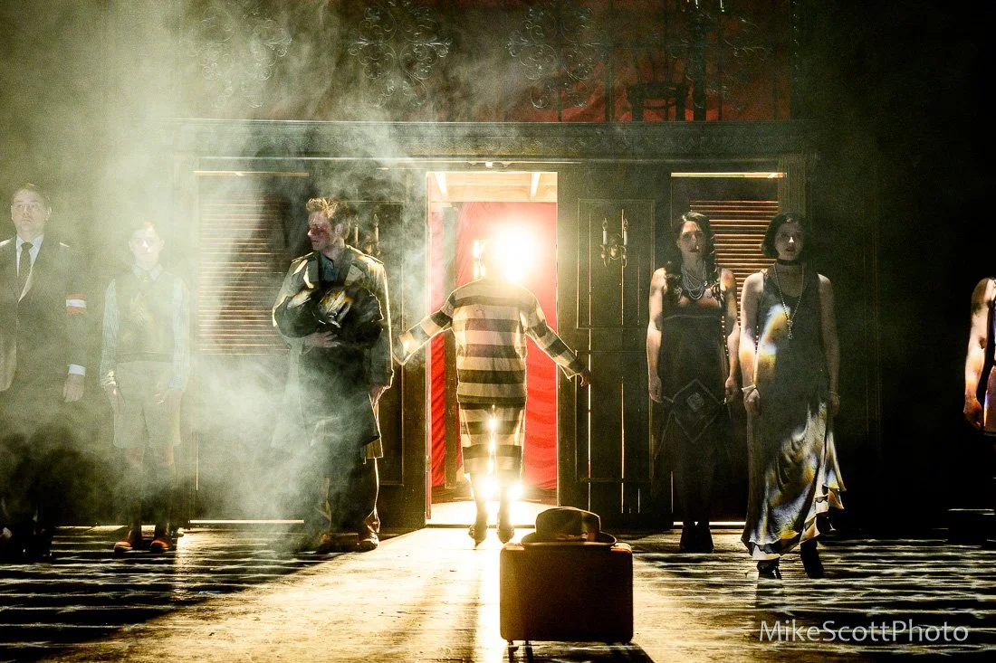 People standing in front of a lit doorway with smoke and bright backlight, some with masks or makeup, others dressed formally or in costumes, on a dark stage.