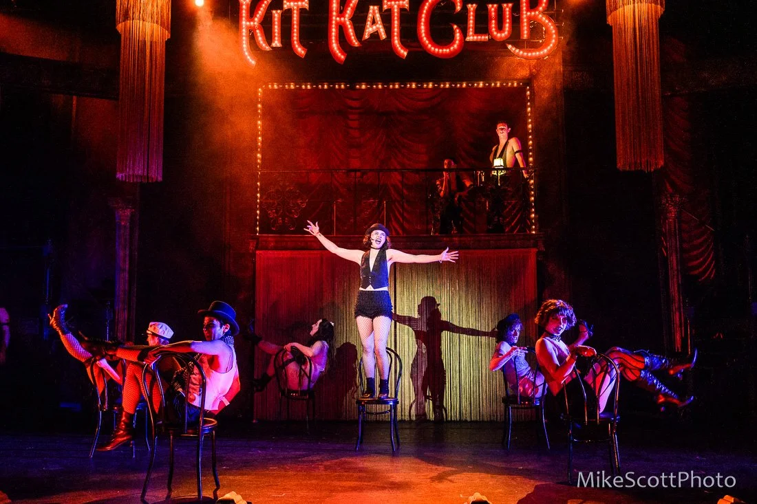 Performers in vintage costumes on stage at Kit Kat Club, with neon sign overhead, theatrical lighting, and red curtains in the background.