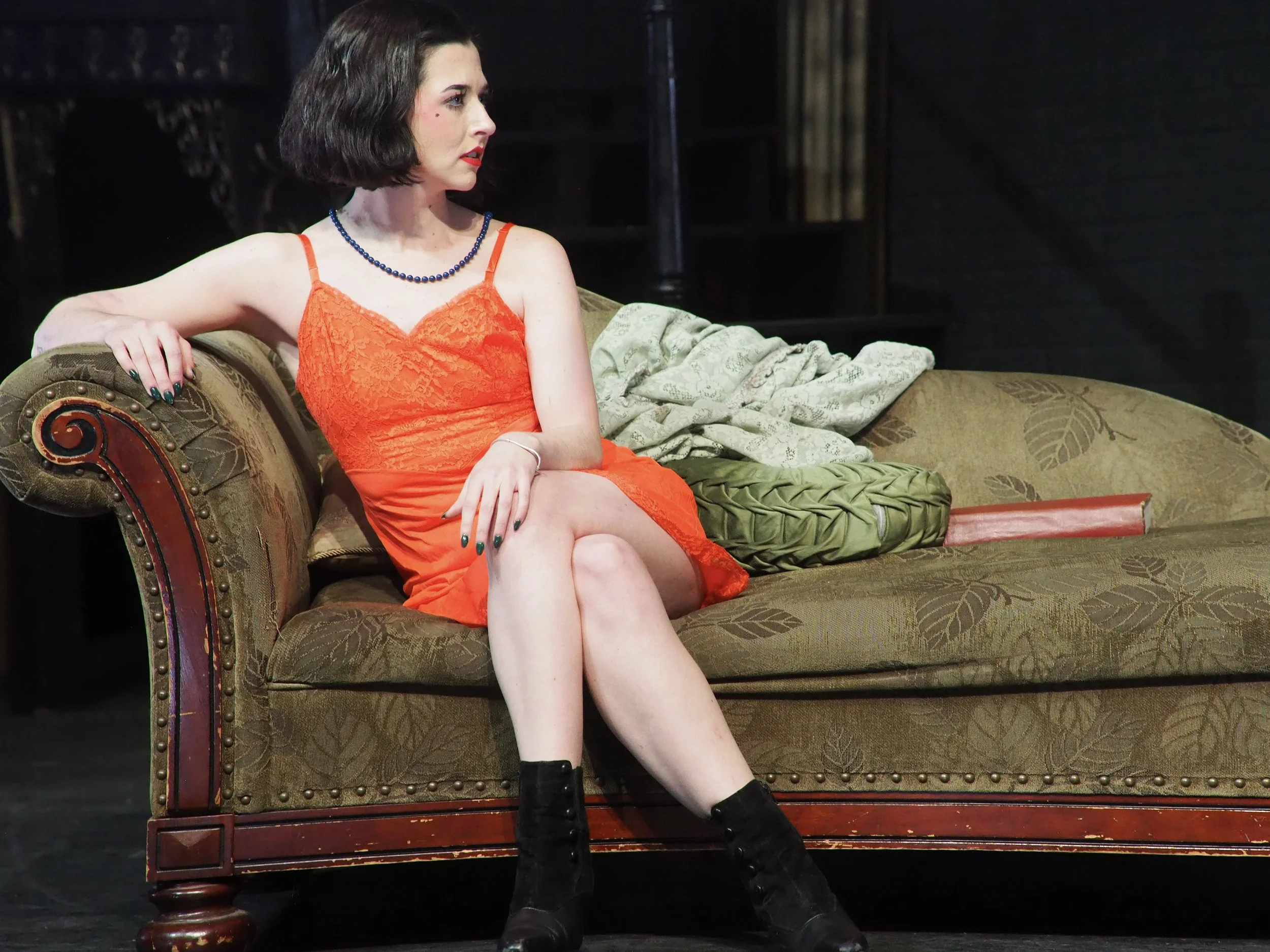A woman with short dark hair, red lipstick, and wearing an orange dress with lace top and spaghetti straps, sits on a vintage-style green sofa with a patterned pillow, looking to her left.