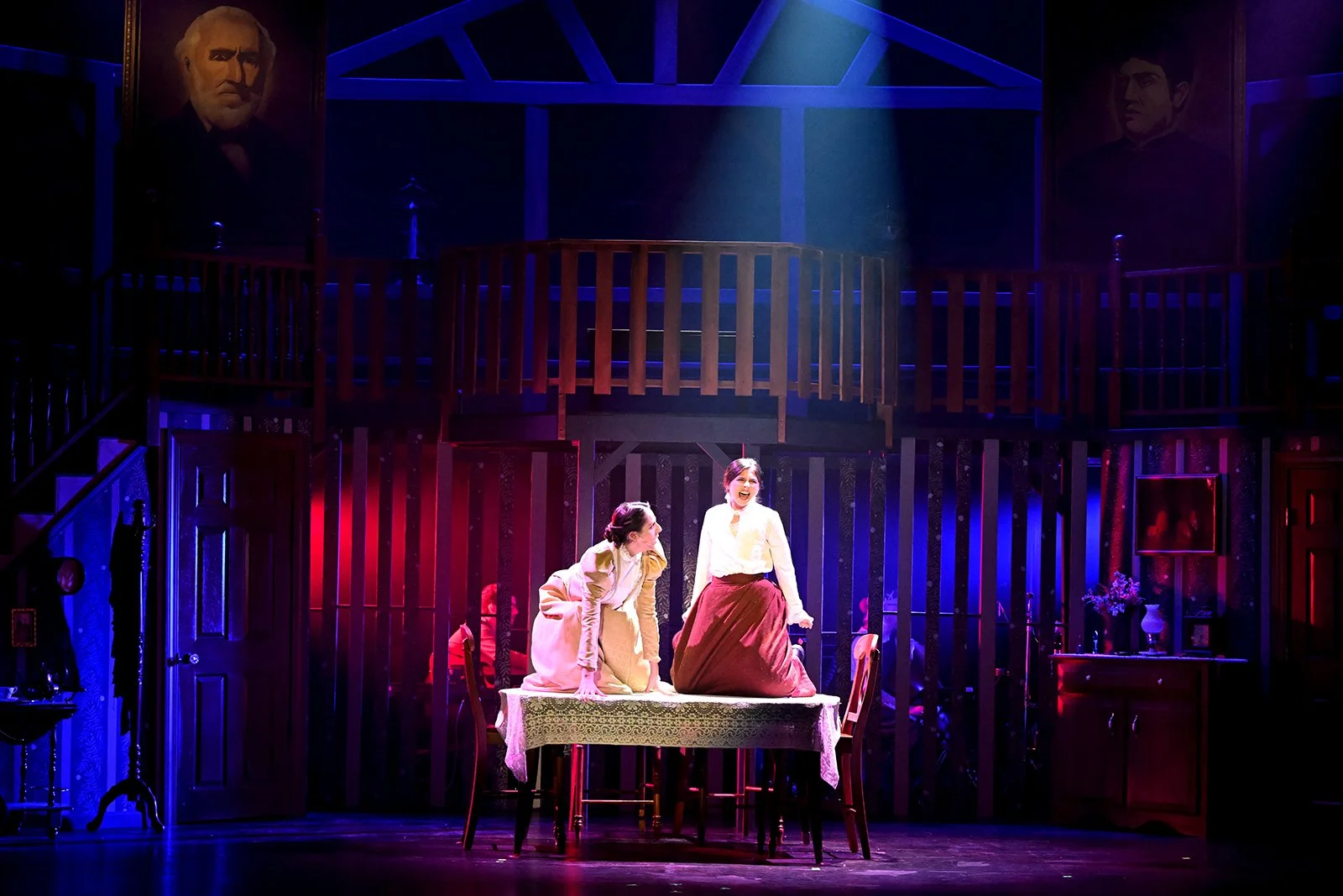Two women in period costumes are on a stage, one standing and smiling, the other kneeling on a table, with a backdrop of a wooden balcony and portrait paintings, under dramatic stage lighting.