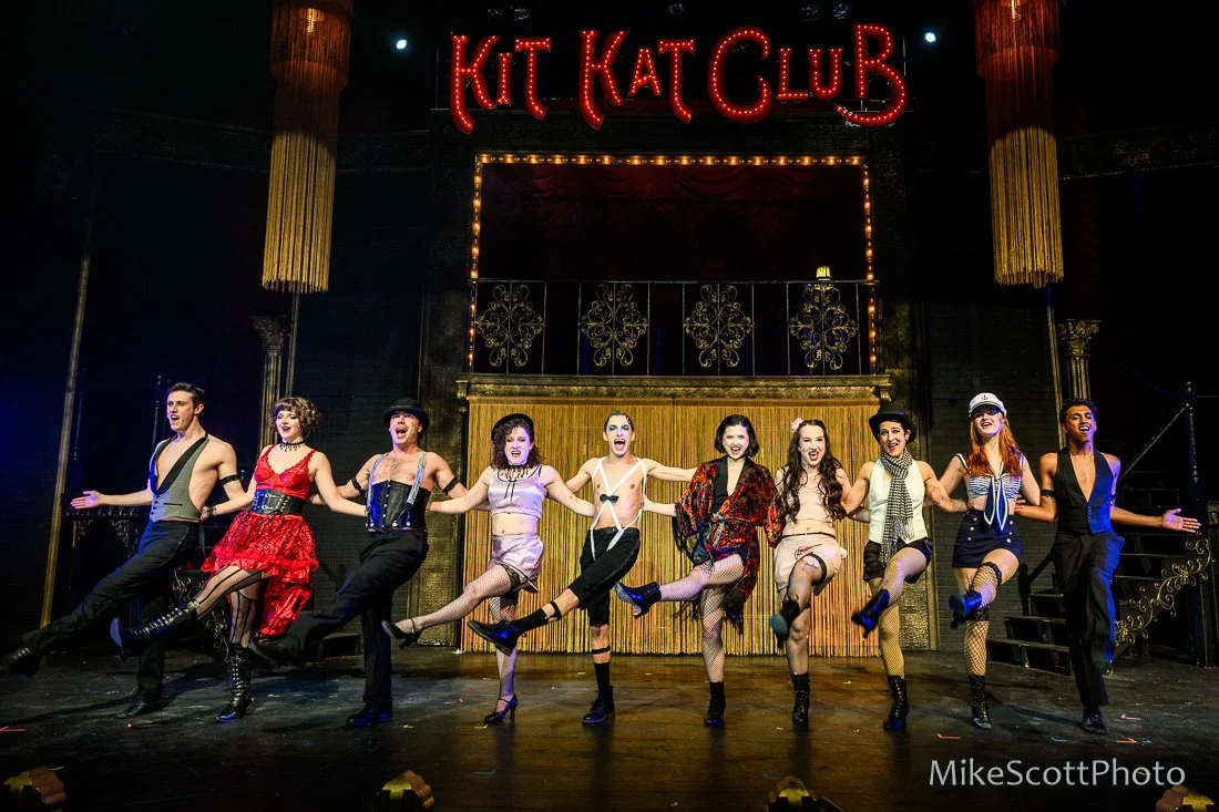 A group of performers on stage at Kit Kat Club, holding hands, mid-dance, with a large sign above that reads 'KIt Kat Club' and stage lighting.
