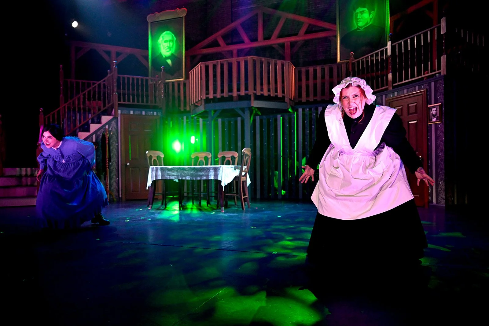 Two women performing on stage with wooden staircase and balcony in the background, portraits hanging on the wall, and a table with chairs in the center. Stage lighting is colorful, with green lights highlighting parts of the scene. One woman is dressed in a black dress with a white apron and bonnet, and the other in a purple dress. Both women are expressive, engaging in their performance.