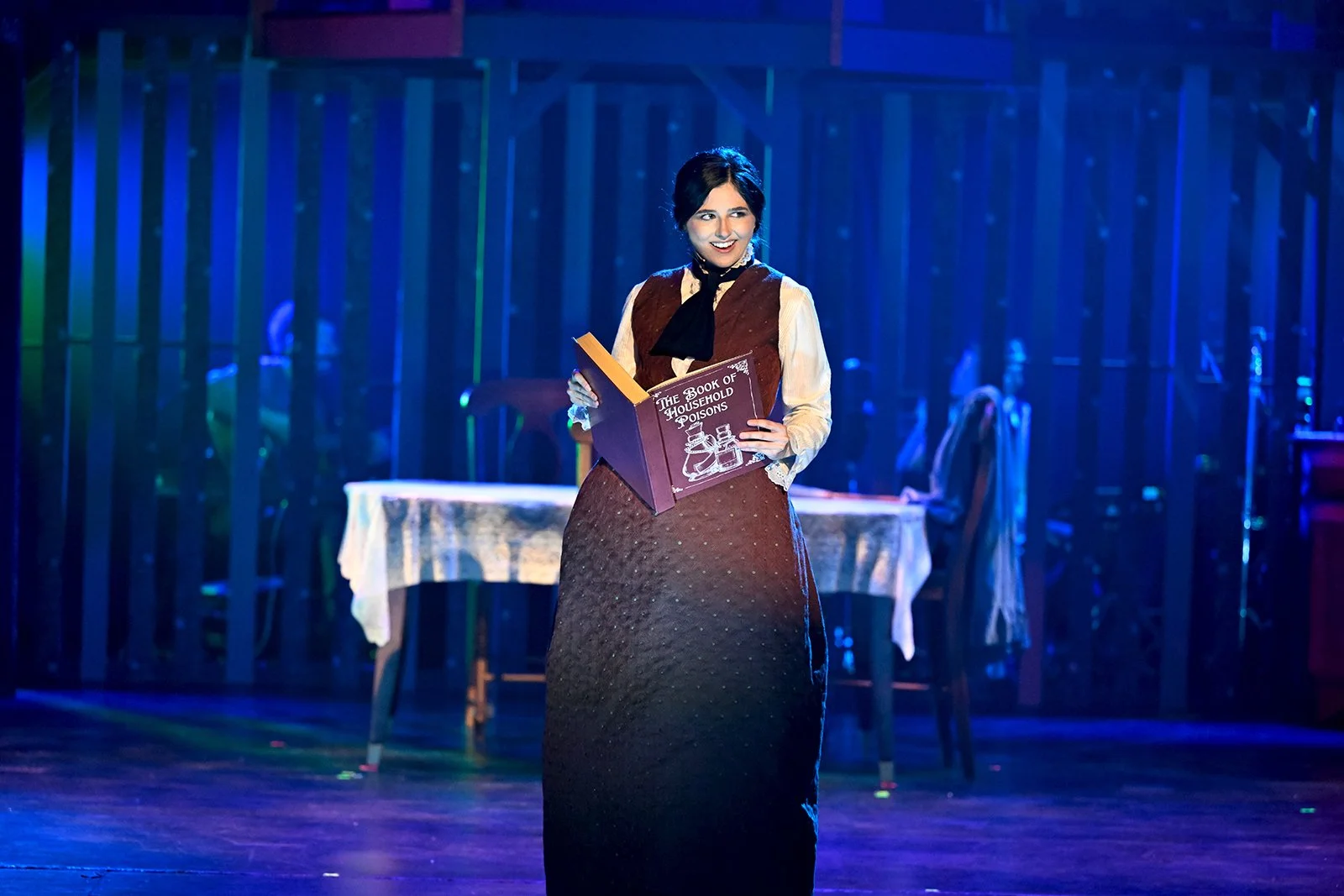 Woman on stage holding a large book titled 'The Book of Household Poisons,' with a table and chairs in the background, stage lighting, and a wooden floor.