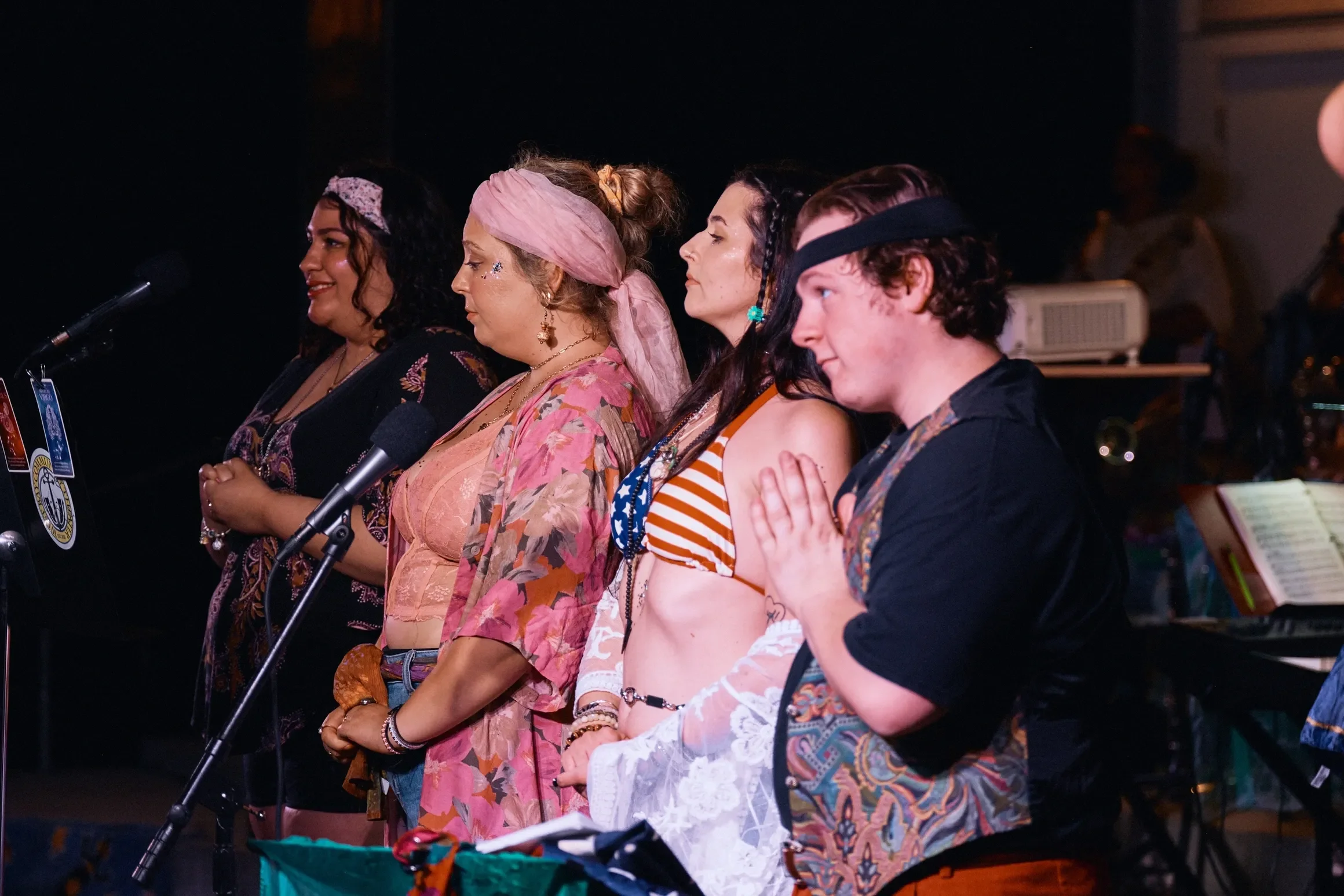 Five people standing in a row with their eyes closed, hands over their hearts, near microphones on a stage.