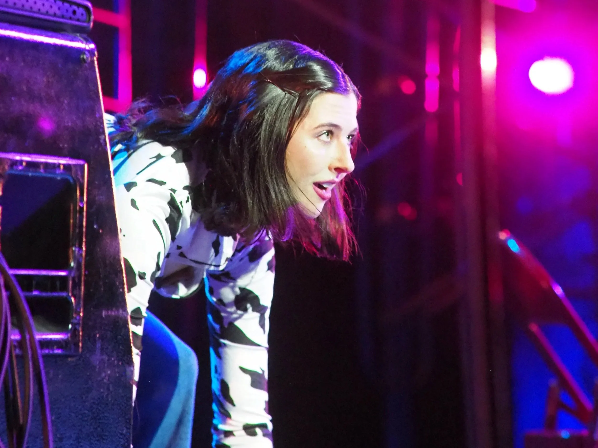 A woman with shoulder-length brown hair leaning over a speakers at a concert with pink and purple stage lights in the background.
