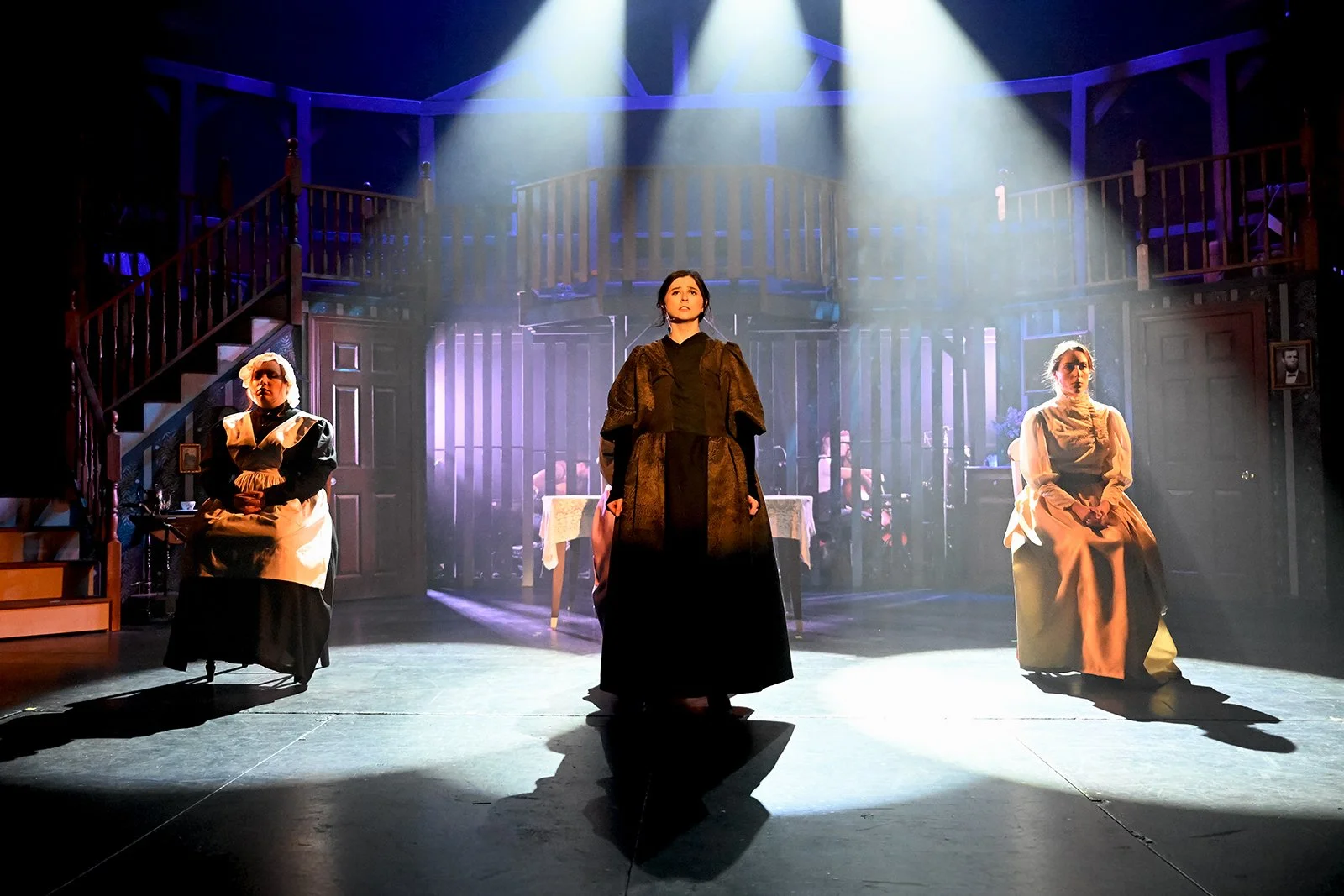 Three women are sitting and standing on a stage with dramatic lighting, a wooden staircase, and a background resembling a house interior, during a theatrical performance.