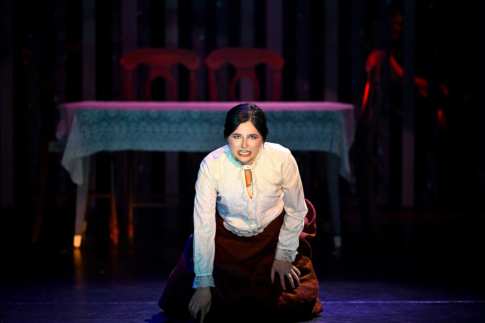 A woman with dark hair kneels on stage in a theatrical performance, wearing a cream blouse and a dark skirt, with a distressed expression, in front of a table with a tablecloth and two chairs in a dimly lit setting.