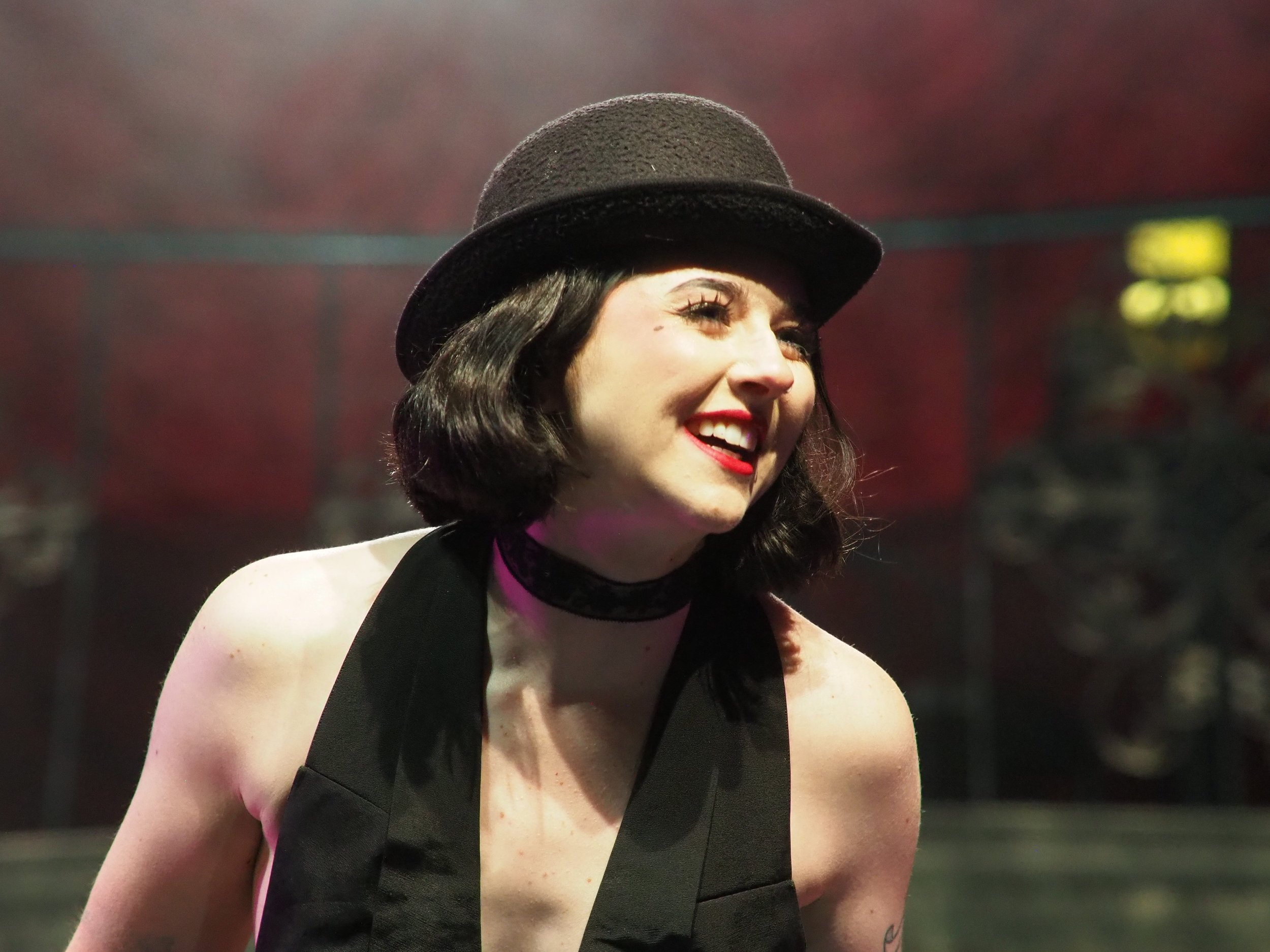 A woman smiling, wearing a black hat, black sleeveless top, and a choker, with a blurred stage with speakers in the background.