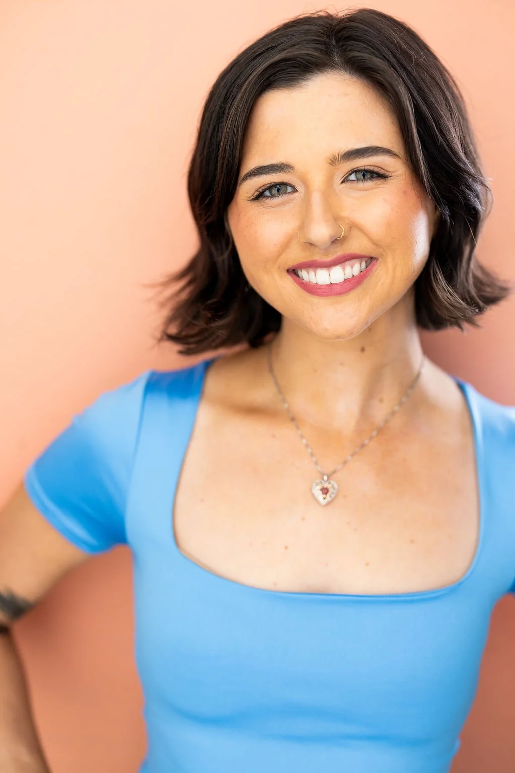 A woman with short dark brown hair, blue eyes, and a nose ring, smiling in front of a peach-colored background, wearing a blue top and a necklace with a heart-shaped pendant.