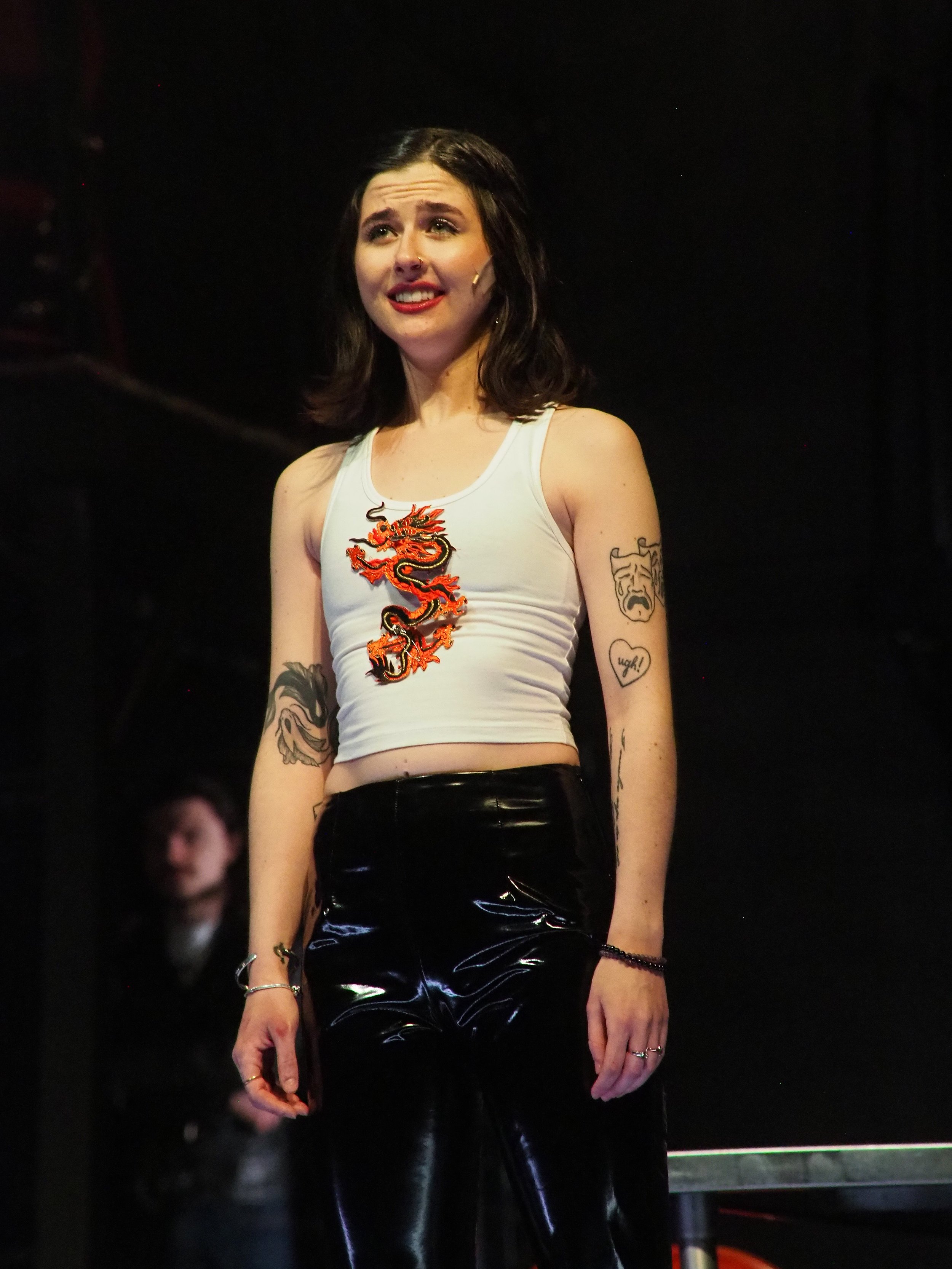 A young woman with dark hair standing on a stage, wearing a white crop top with a dragon design and black shiny pants. She has several tattoos on her arms and is smiling with a slightly anxious expression.